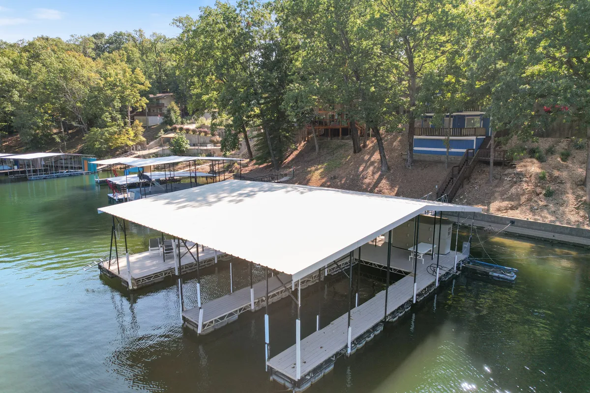 A tranquil scene featuring a dock extending over calm lake waters, surrounded by trees and with houses perched on the hillside. The inviting water, accessible dock, and peaceful setting highlight the lakeside lifestyle and opportunities for boating, swimming, and fishing.
