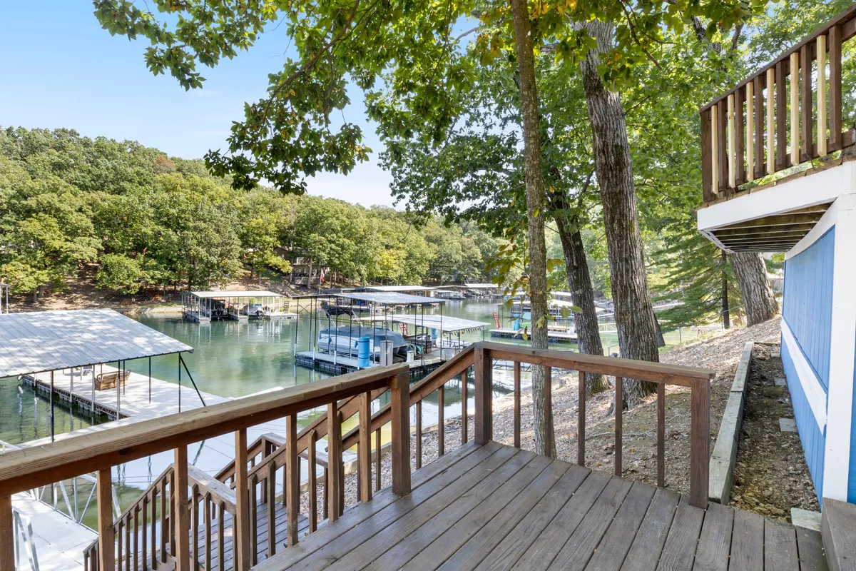 A scenic, sunlit wooden deck stretches out towards the water, with a private dock and boat slips visible. Lush greenery surrounds the waterfront, and boats are moored at the dock, illustrating direct lake access and leisurely lakeside living.