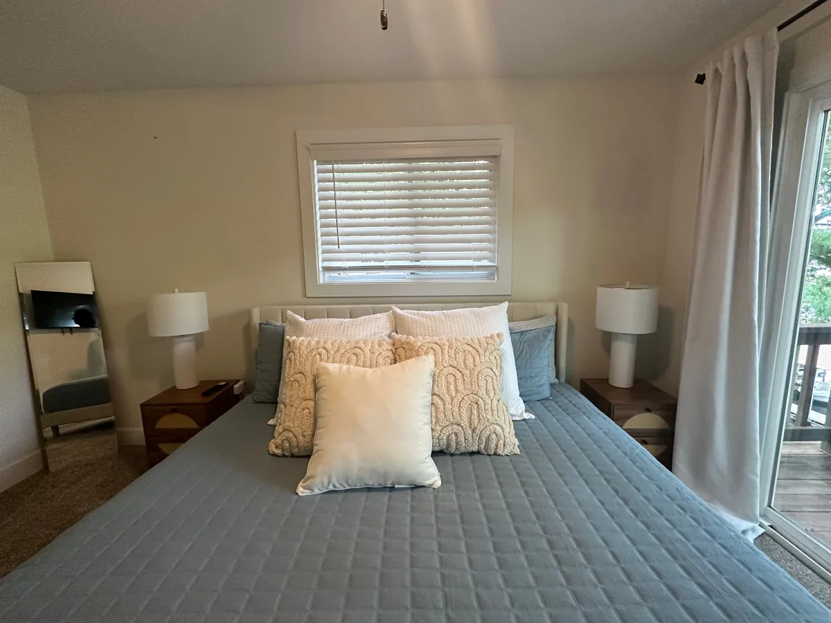 A tranquil bedroom featuring a cozy bed with decorative pillows, a nightstand with a lamp, and a mirror. The space is bathed in natural light, inviting relaxation and moments of quiet comfort.