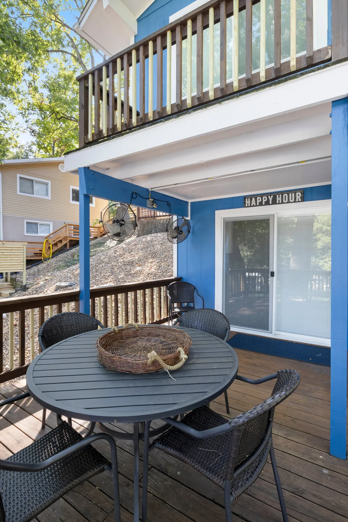 An inviting outdoor seating area featuring a round dining table, decorative basket, and a cheerful sign reading 'HAPPY HOUR.' The setting is designed for al fresco dining and socializing, with ample space for refreshments and gatherings under the open sky.