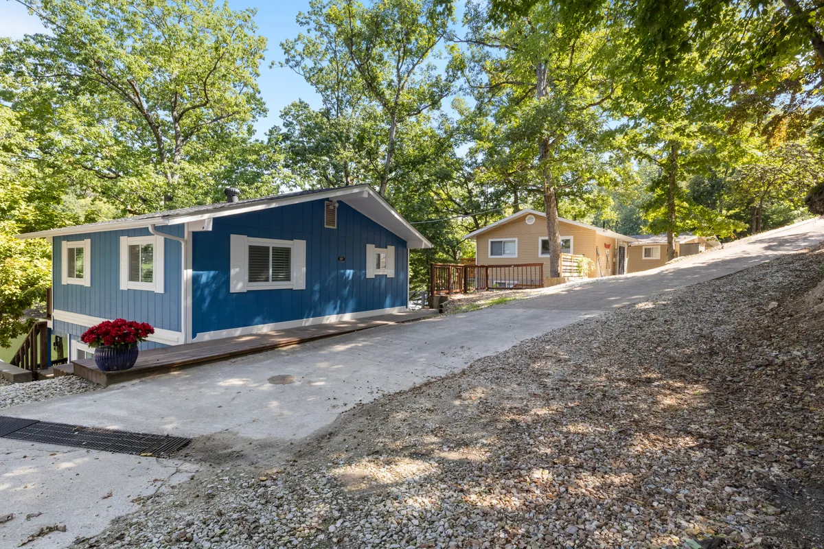 A charming blue and yellow cabin sit beneath a canopy of trees, with a gravel path and a flower pot inviting guests to explore or gather outside. The scene feels cozy and welcoming, perfect for family groups arriving or enjoying the outdoors together.
