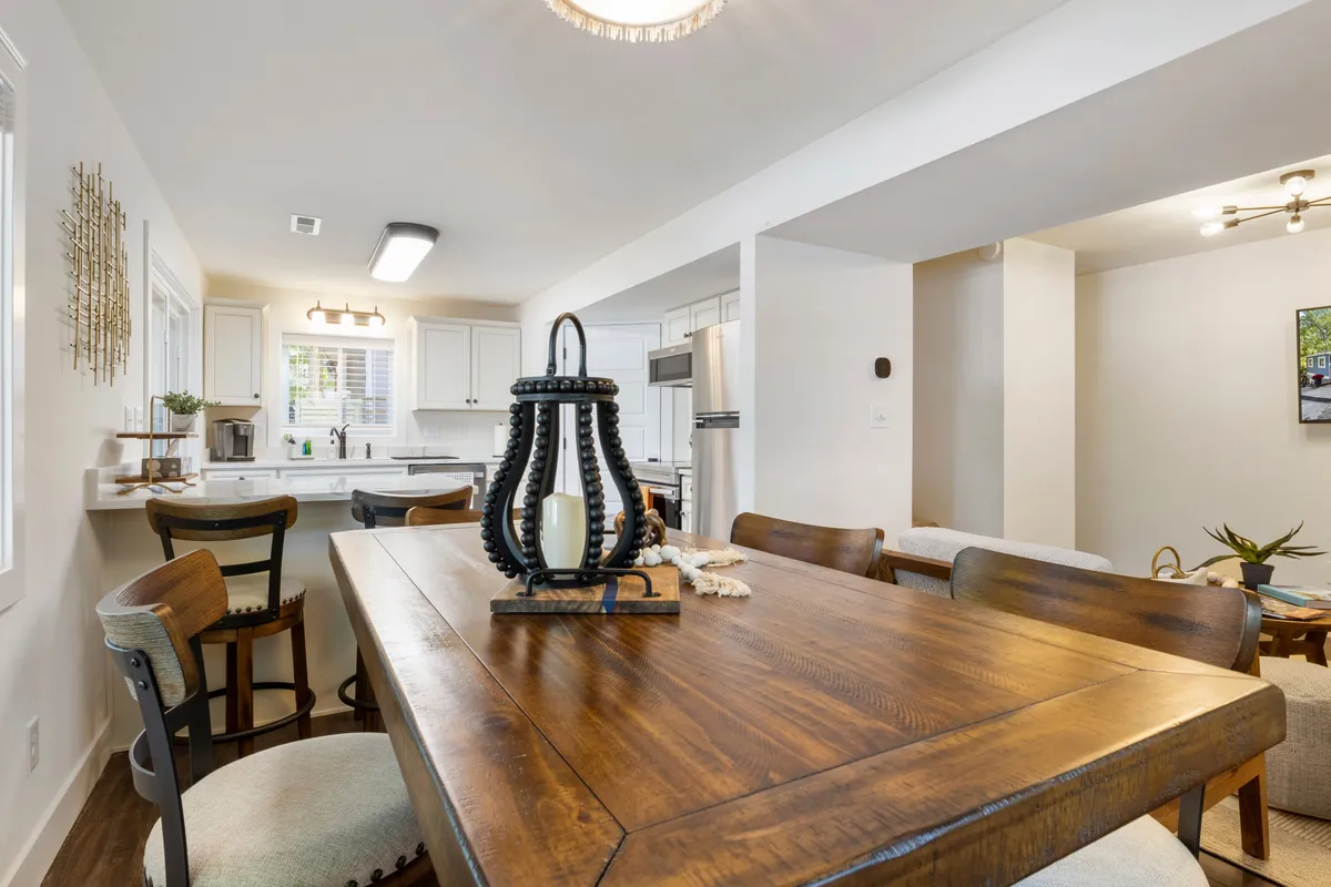 A warm dining area with a large wooden table, a decorative lantern centerpiece, and the kitchen visible in the background. The space is clearly set up for shared meals, entertaining guests, and creating family traditions, with ample seating provided.