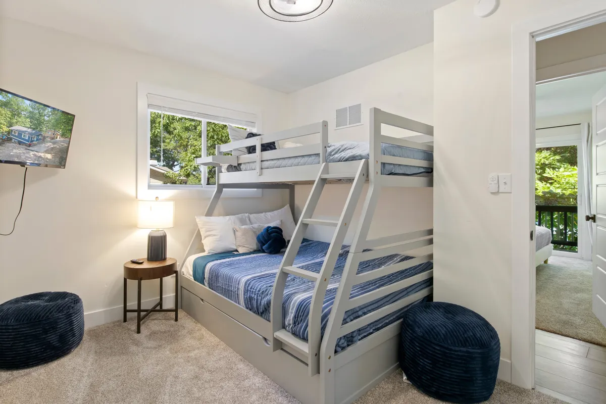 A welcoming bedroom featuring a sturdy bunk bed setup, with a TV on the wall, table lamp, and blue poufs. The design is playful yet safe, making it ideal for children or younger guests, and speaks directly to flexible sleeping arrangements for families.
