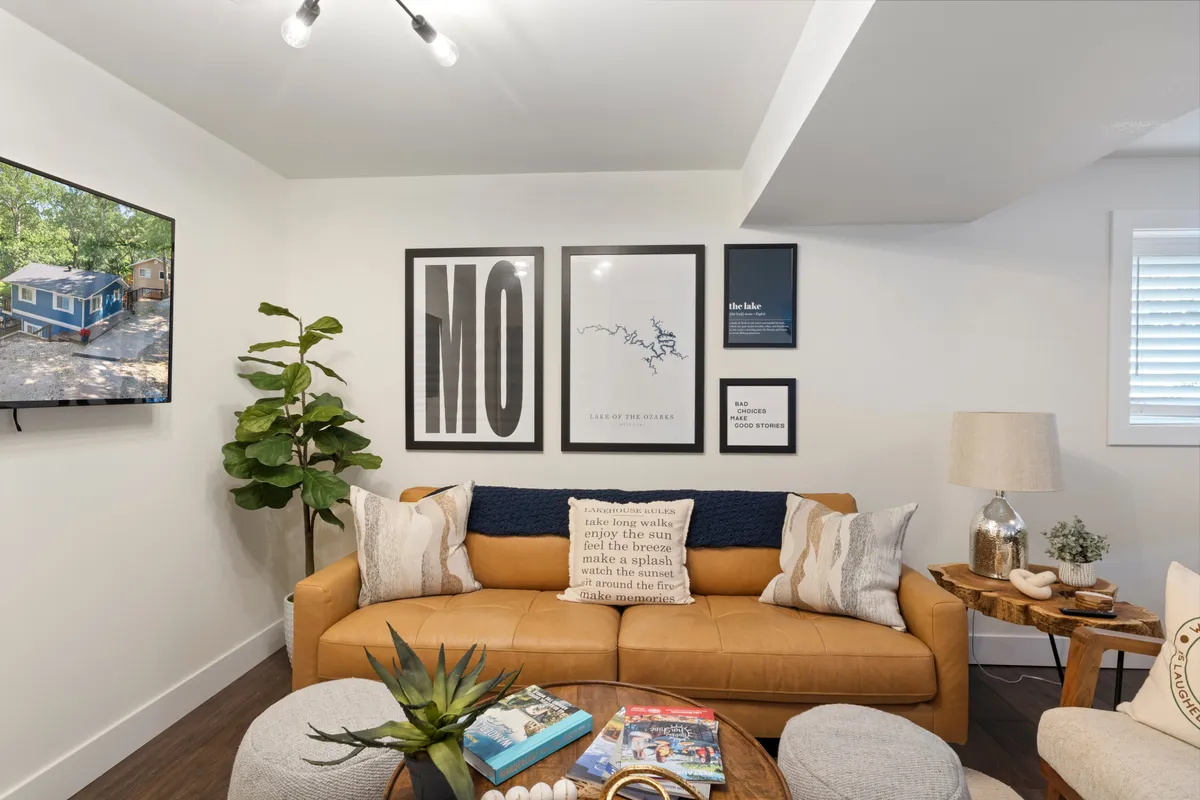The hero image showcases a stylish, cozy living room with a plush leather couch, inviting coffee table, tasteful wall art, a large TV for entertainment, and a leafy potted plant adding homely warmth. The space is bathed in comfortable lighting, emphasizing the room’s welcoming vibe and suitability for gatherings or unwinding after a day by the lake.